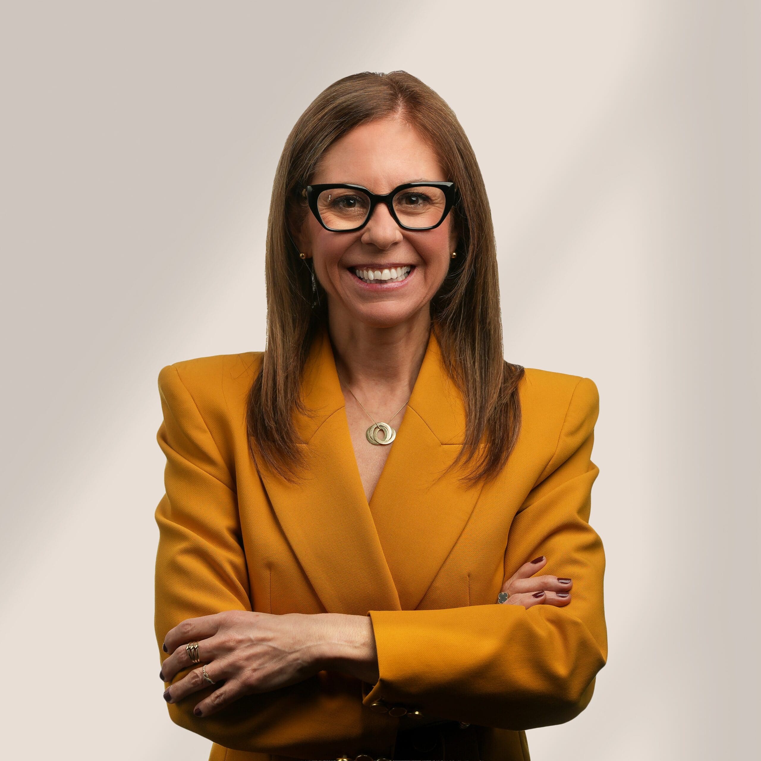 Une femme aux cheveux bruns raides et aux lunettes sourit avec assurance. Elle porte un blazer jaune moutarde et un collier, et se tient debout, les bras croisés, sur un fond clair.