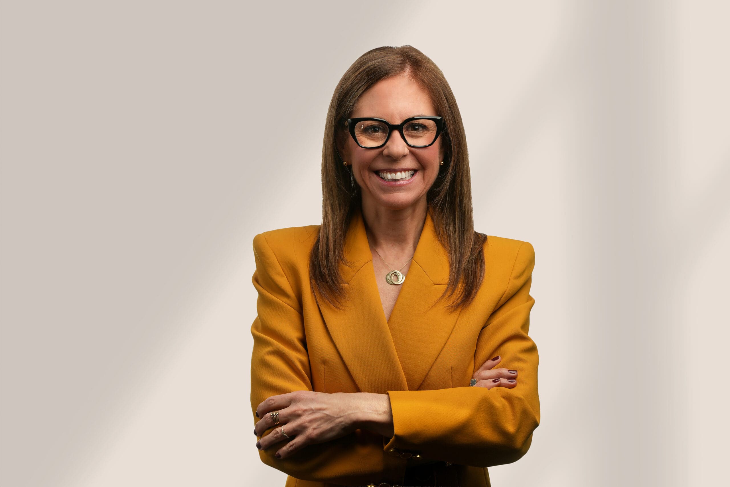 Une femme aux cheveux bruns raides et aux lunettes noires sourit avec assurance, les bras croisés, vêtue d'un blazer jaune moutarde, sur un fond clair et légèrement ombragé.