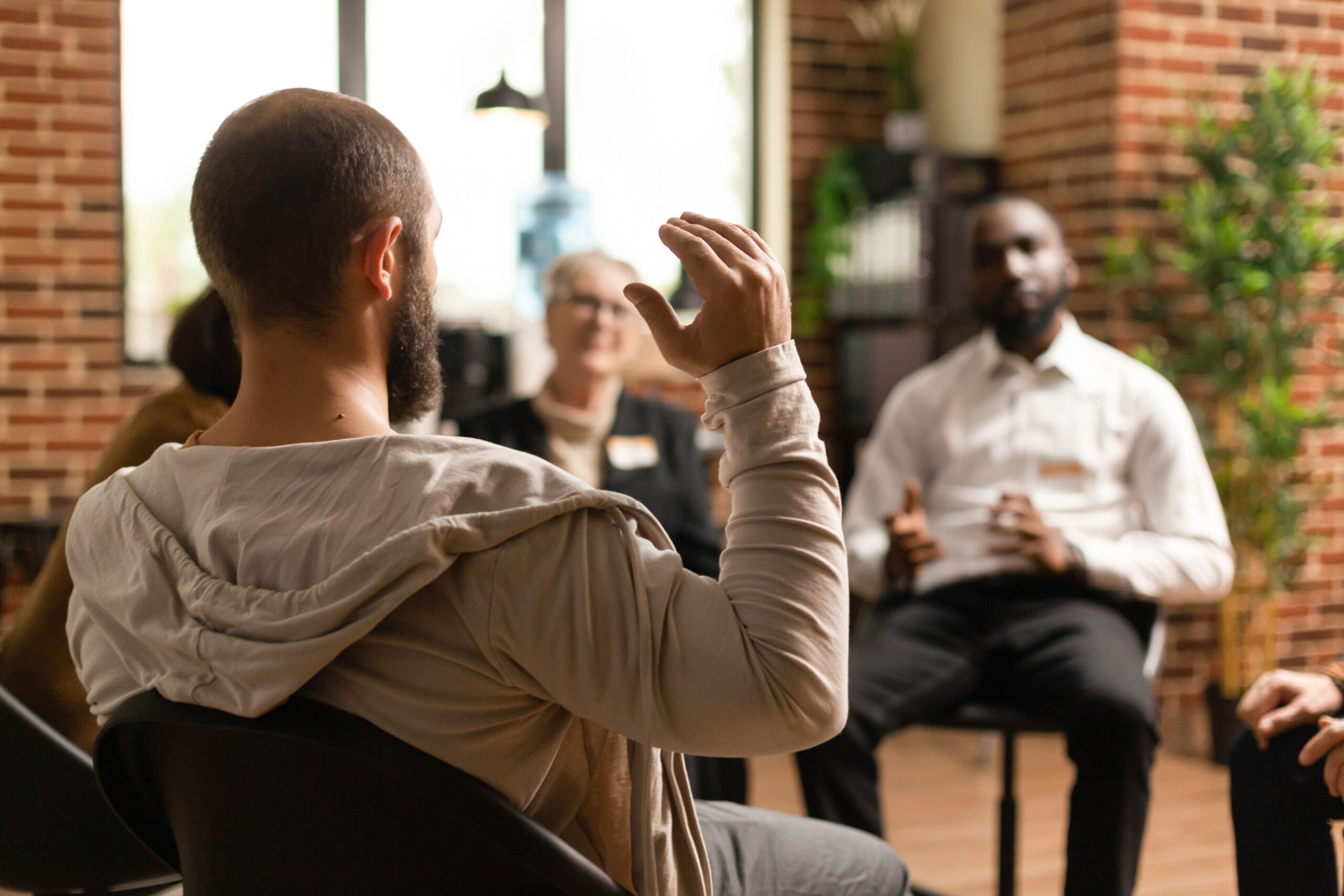 Un groupe de personnes est assis en cercle à l'intérieur, engagé dans une discussion. Une personne au premier plan lève la main tandis que d'autres écoutent attentivement. Le décor est constitué de murs en briques et de grandes fenêtres.