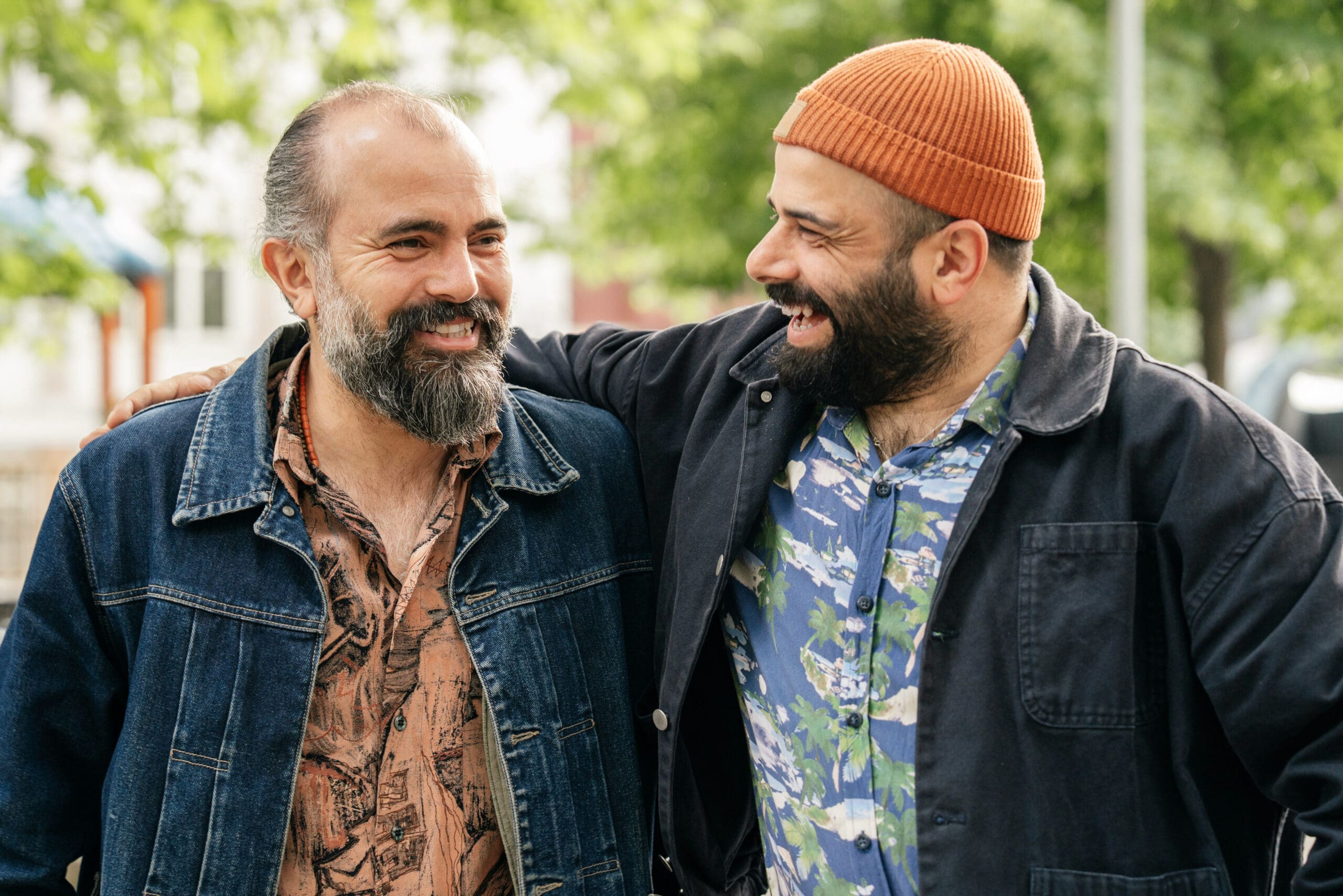 Deux hommes barbus sourient et rient ensemble en plein air. L'un d'eux a le bras autour de l'autre. Tous deux portent des chemises et des vestes à motifs ; l'un d'eux porte un bonnet en tricot orange. Des arbres verts sont flous à l'arrière-plan.
