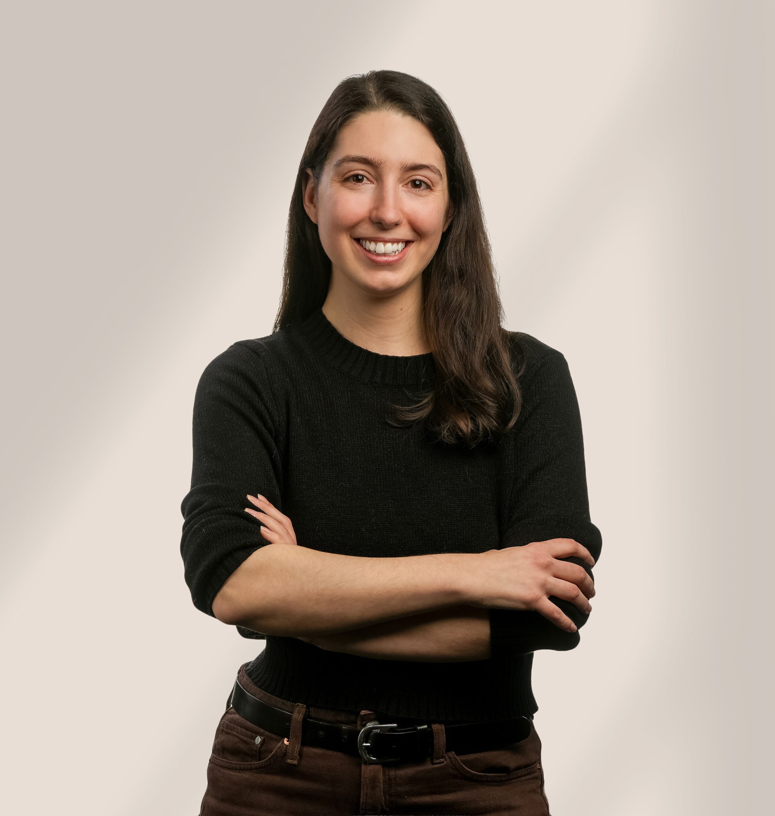 Une femme aux longs cheveux noirs, portant un pull noir et un pantalon foncé, sourit, les bras croisés, sur un fond clair et neutre.
