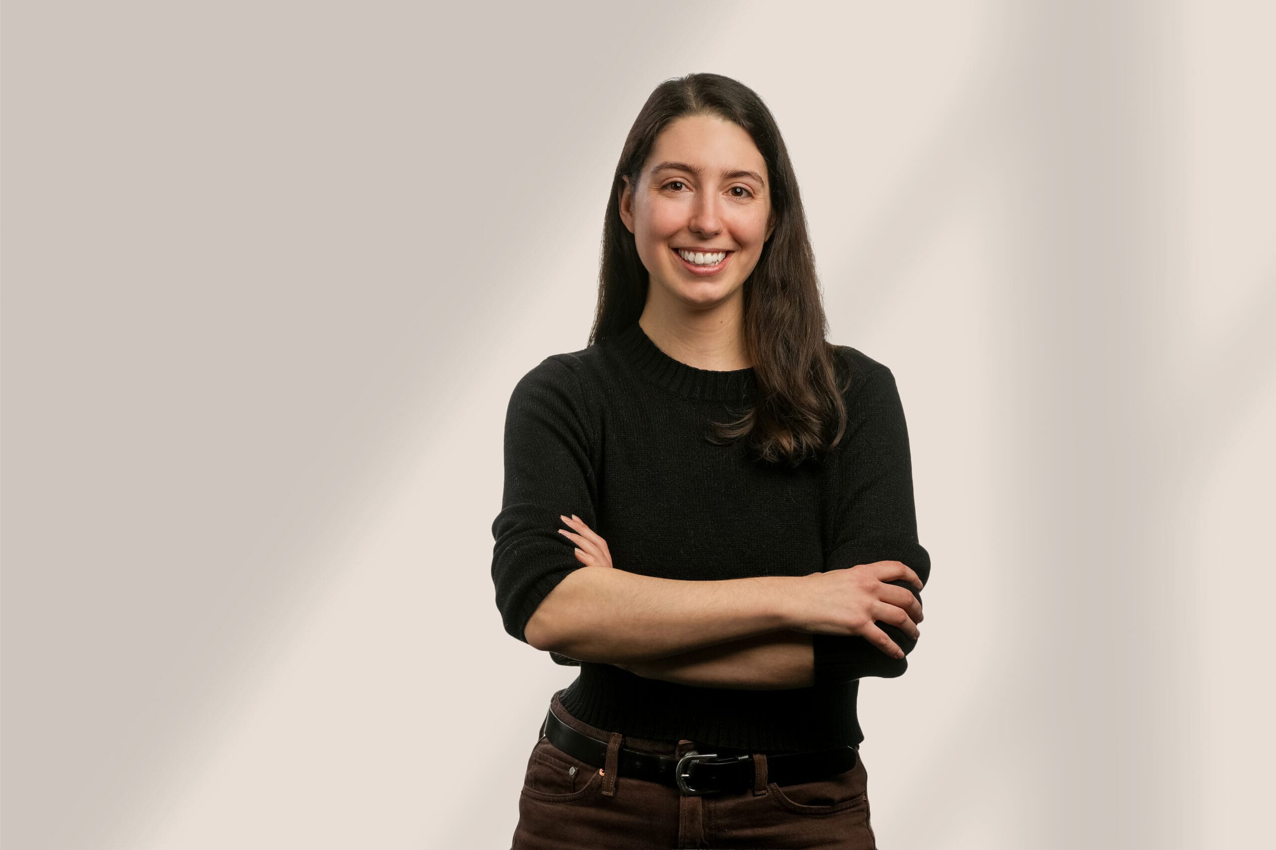 Une femme aux longs cheveux noirs, vêtue d'un pull noir et d'un pantalon marron, se tient debout, souriante, les bras croisés, sur un fond clair et légèrement ombragé.