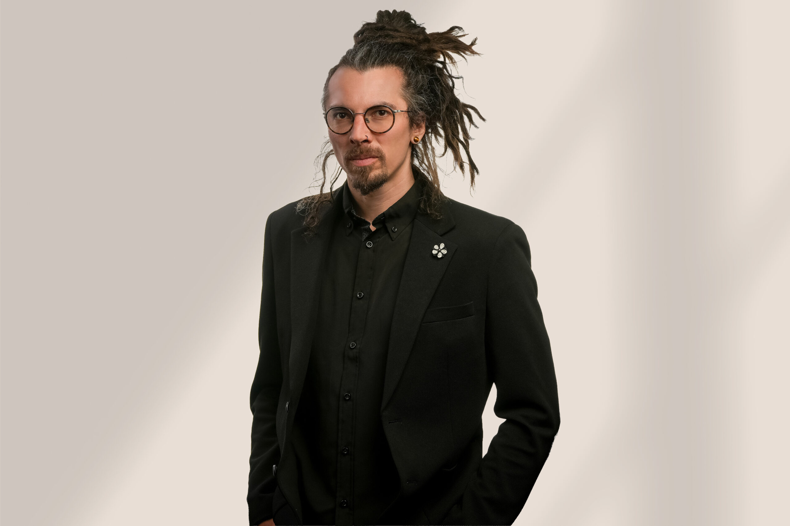 Un homme portant de longues dreadlocks, des lunettes et une barbe, vêtu d'un costume et d'une chemise noirs, se tient debout sur un fond uni de couleur claire, les mains dans les poches.