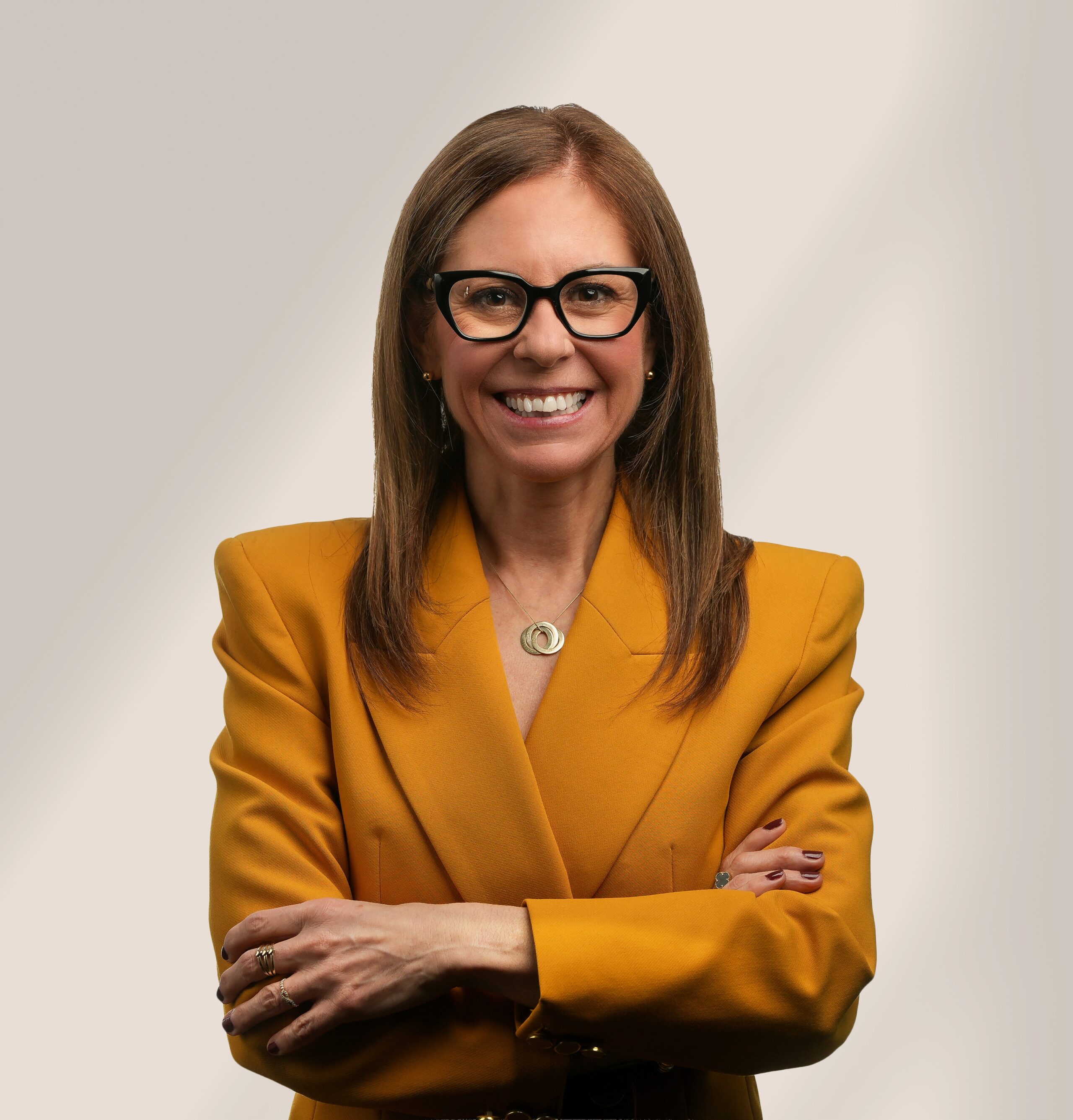 Une femme aux longs cheveux bruns, portant des lunettes noires et un blazer jaune moutarde, sourit avec assurance, les bras croisés, devant un fond neutre.