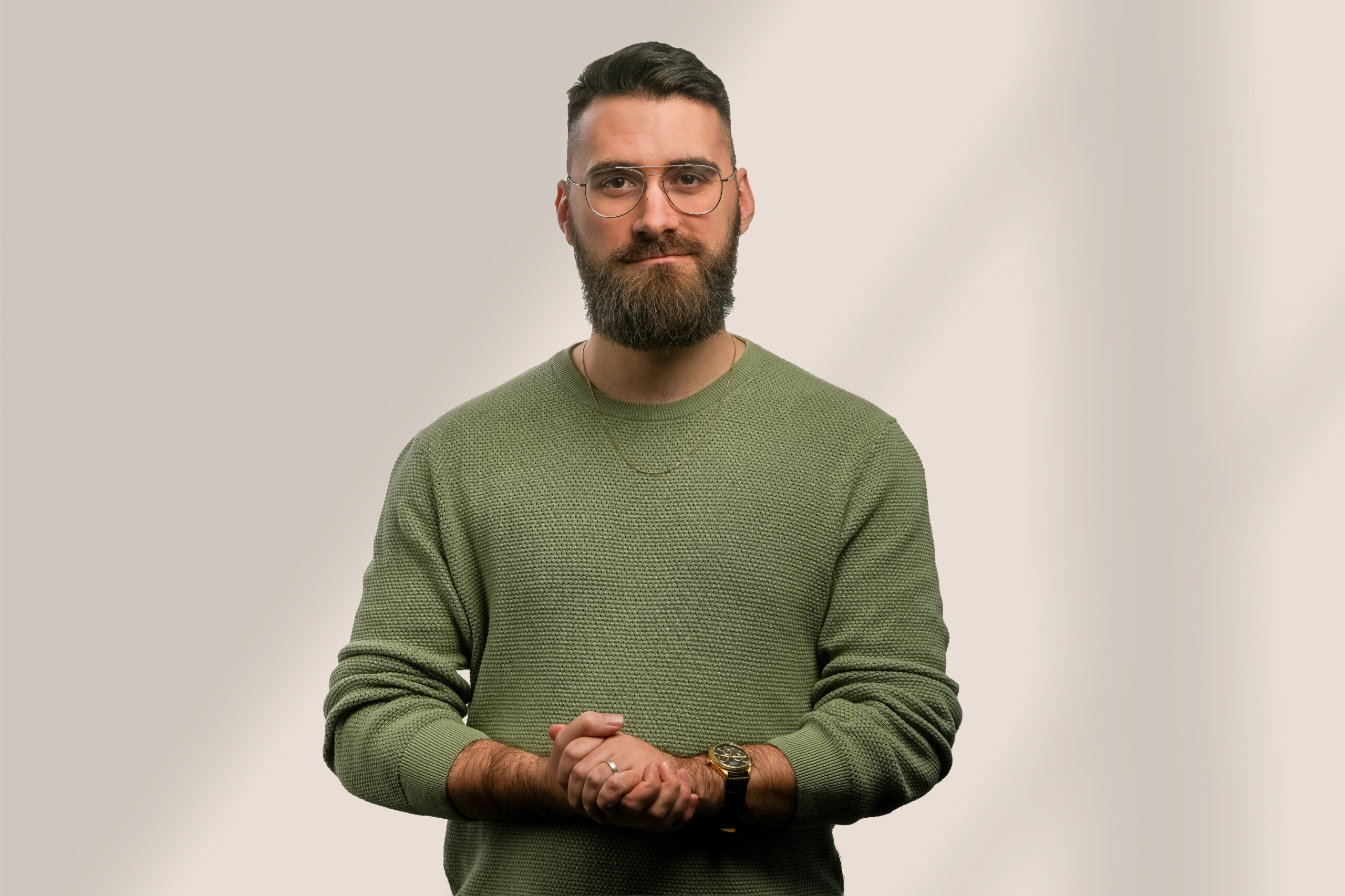 Un homme barbu à lunettes, portant un pull vert et une montre, se tient sur un fond clair et uni, regardant l'appareil photo, les mains jointes devant lui.