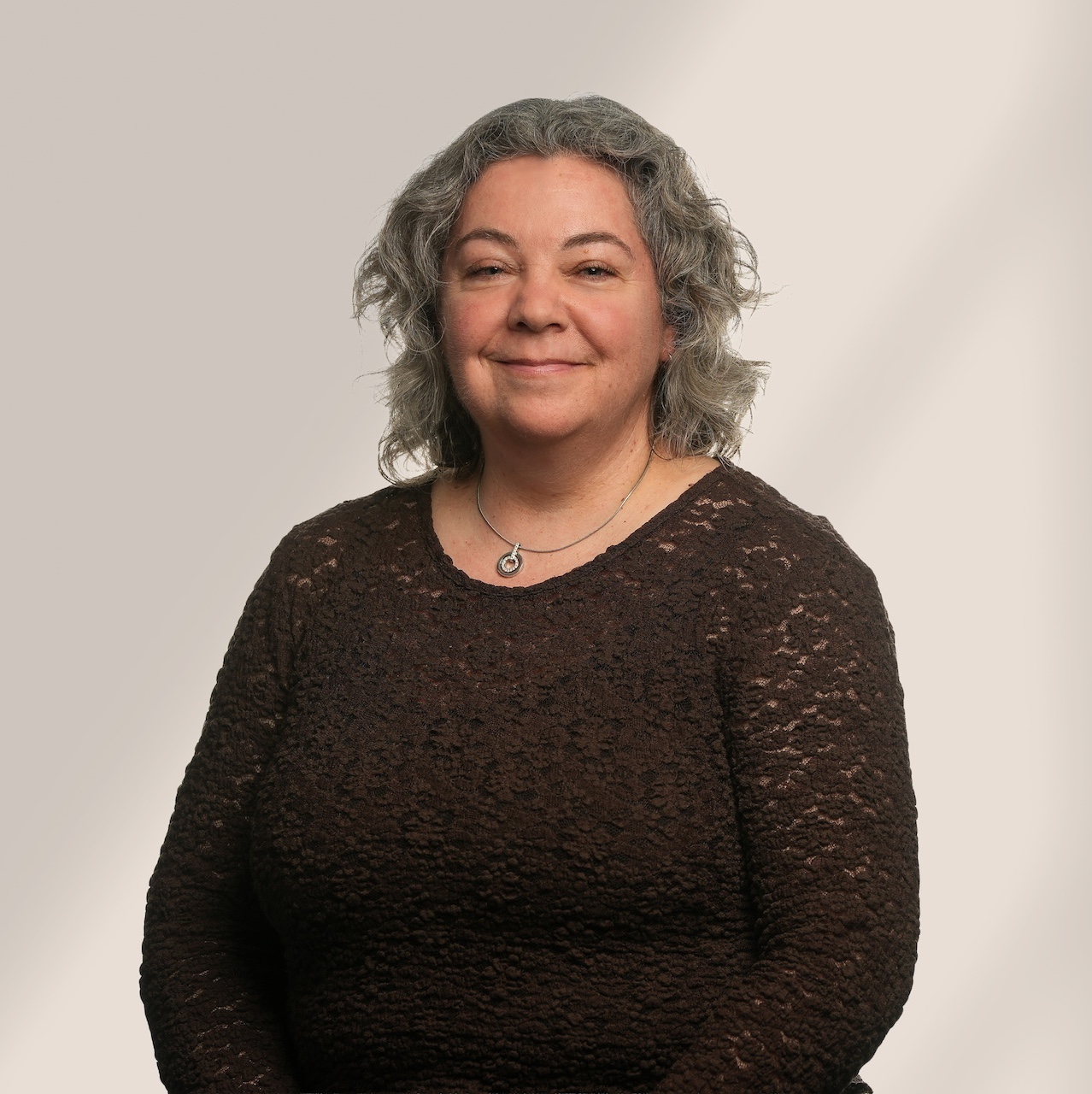 Une femme aux cheveux gris bouclés, portant un haut brun foncé texturé et un collier argenté, est assise et sourit doucement sur un fond gris clair.