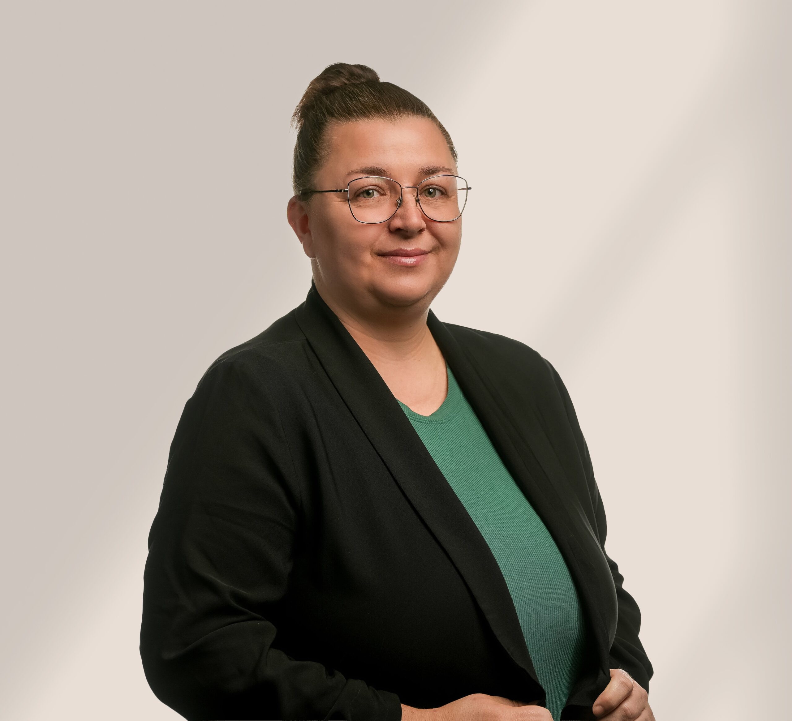 Une personne avec des lunettes et les cheveux en chignon, portant un blazer noir sur une chemise verte, se tient sur un fond beige clair, souriant légèrement à l'appareil photo.