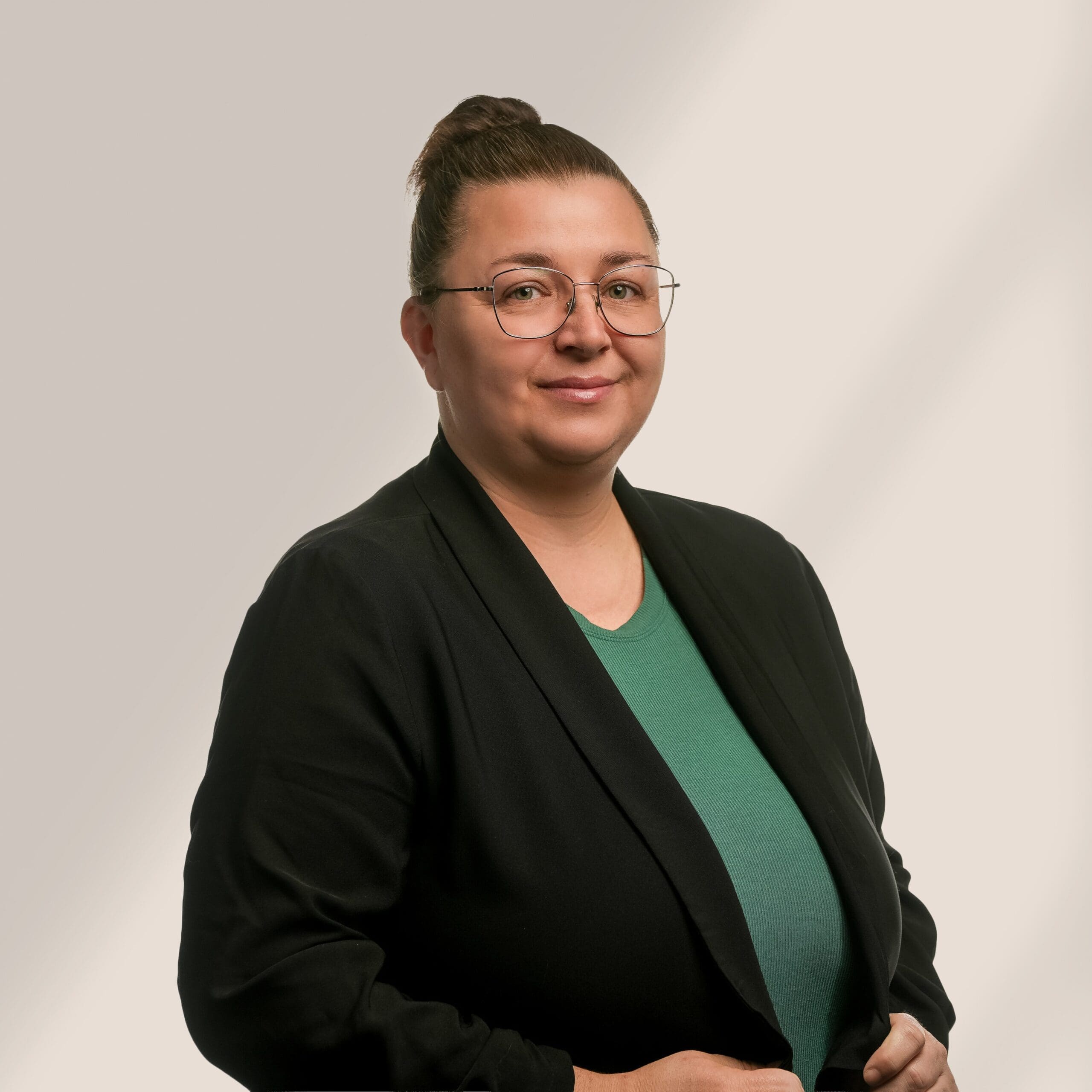 Une personne à lunettes, cheveux bruns en chignon, vêtue d'un haut vert et d'un blazer noir, se tient devant un fond clair uni et sourit légèrement à l'appareil photo.