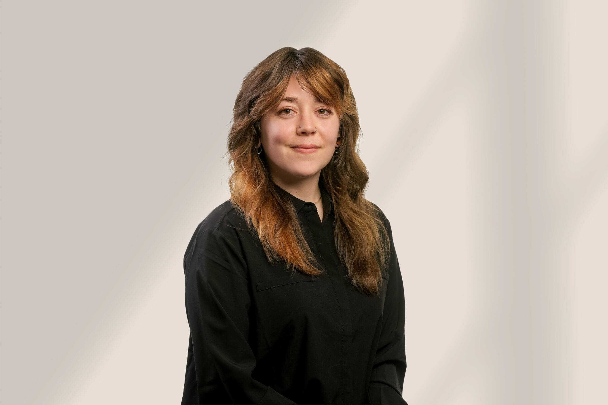 Une femme aux longs cheveux bruns ondulés, vêtue d'une chemise noire, pose devant un fond clair et neutre. Elle fait face à l'appareil photo et sourit doucement.