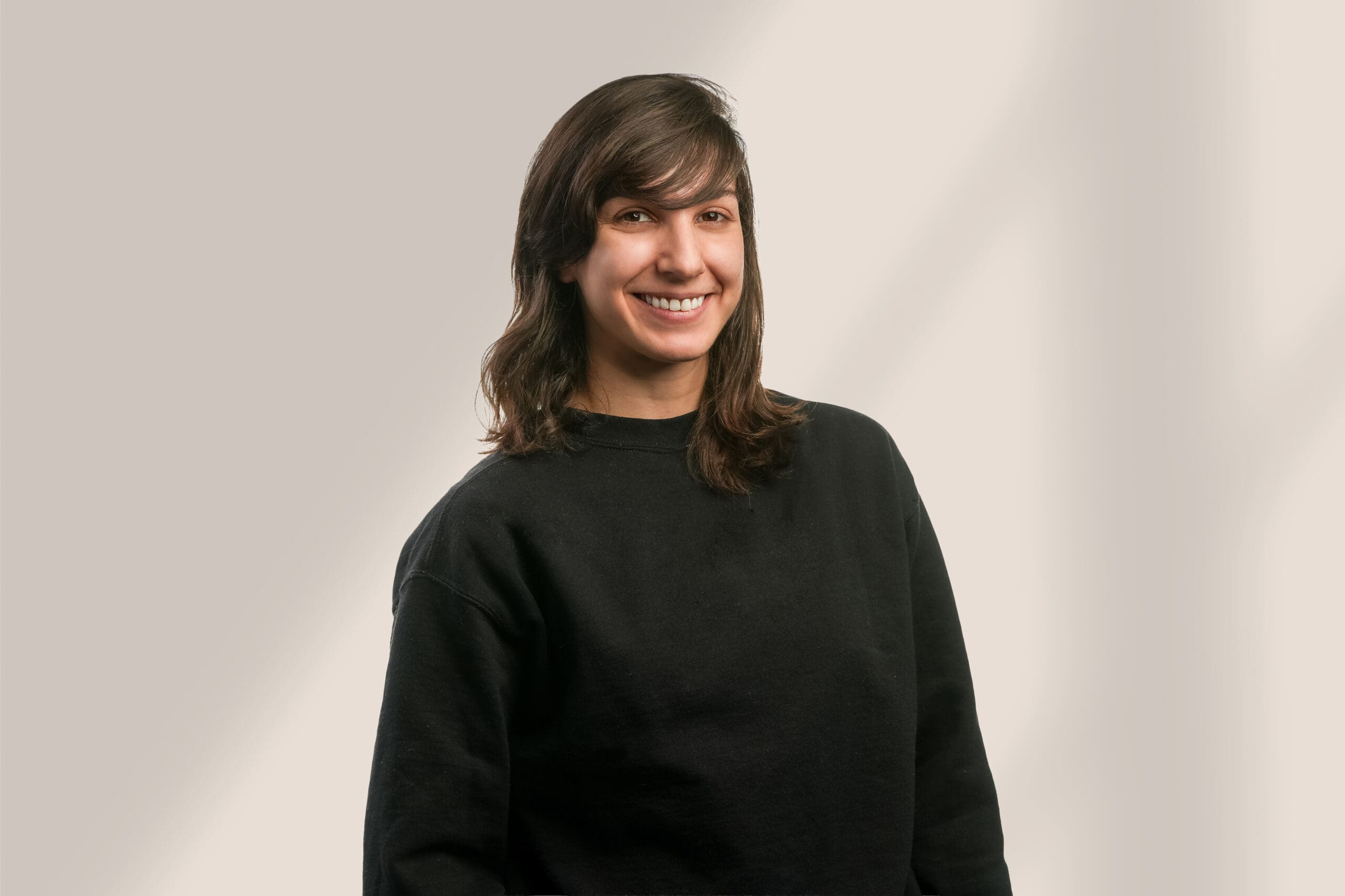 Une jeune personne aux longs cheveux bruns, vêtue d'un pull noir, sourit sur un fond uni de couleur claire.