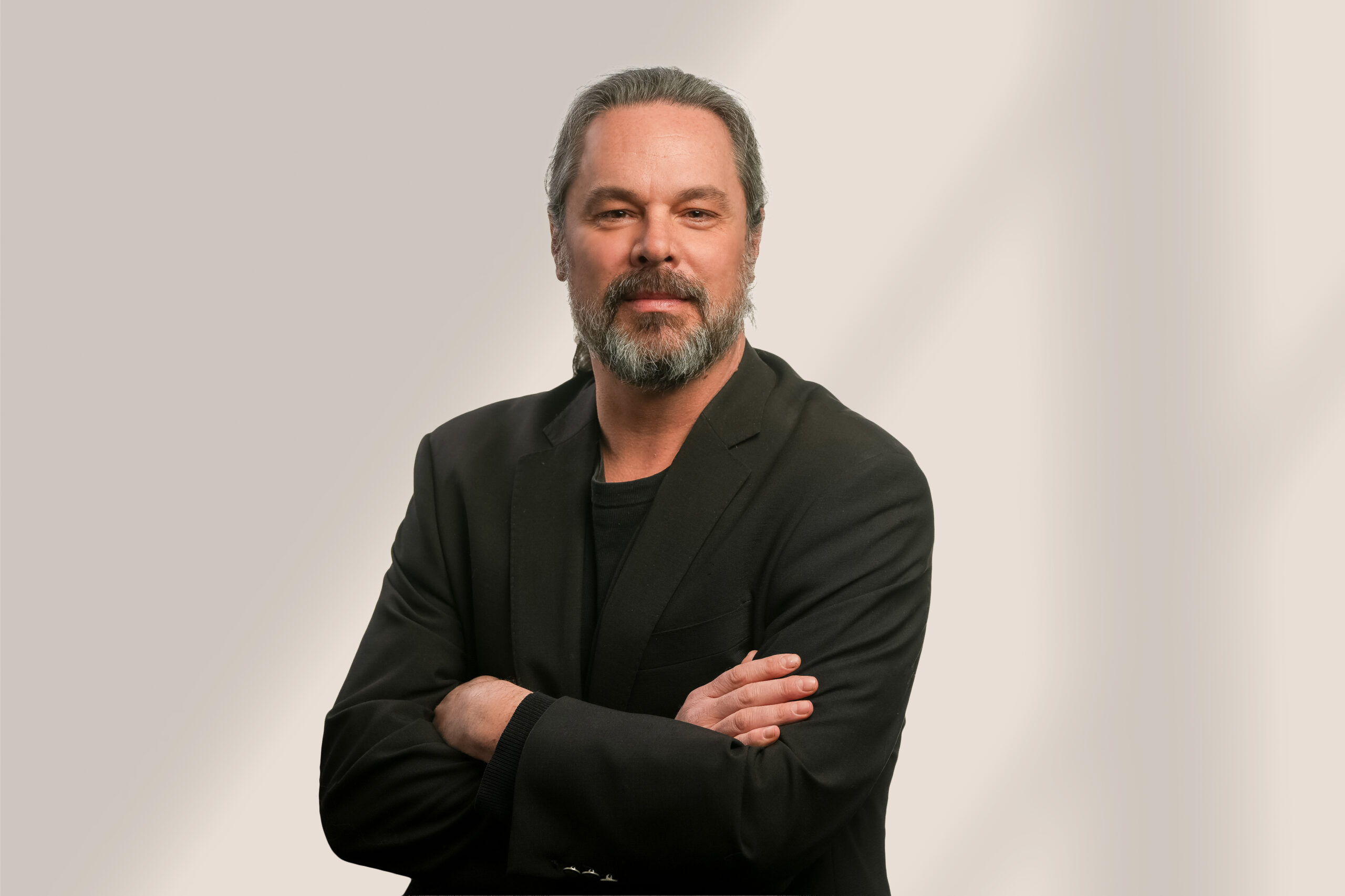 Un homme aux cheveux gris et à la barbe se tient sur un fond clair, vêtu d'un blazer et d'une chemise noirs, les bras croisés, regardant l'appareil photo avec assurance.