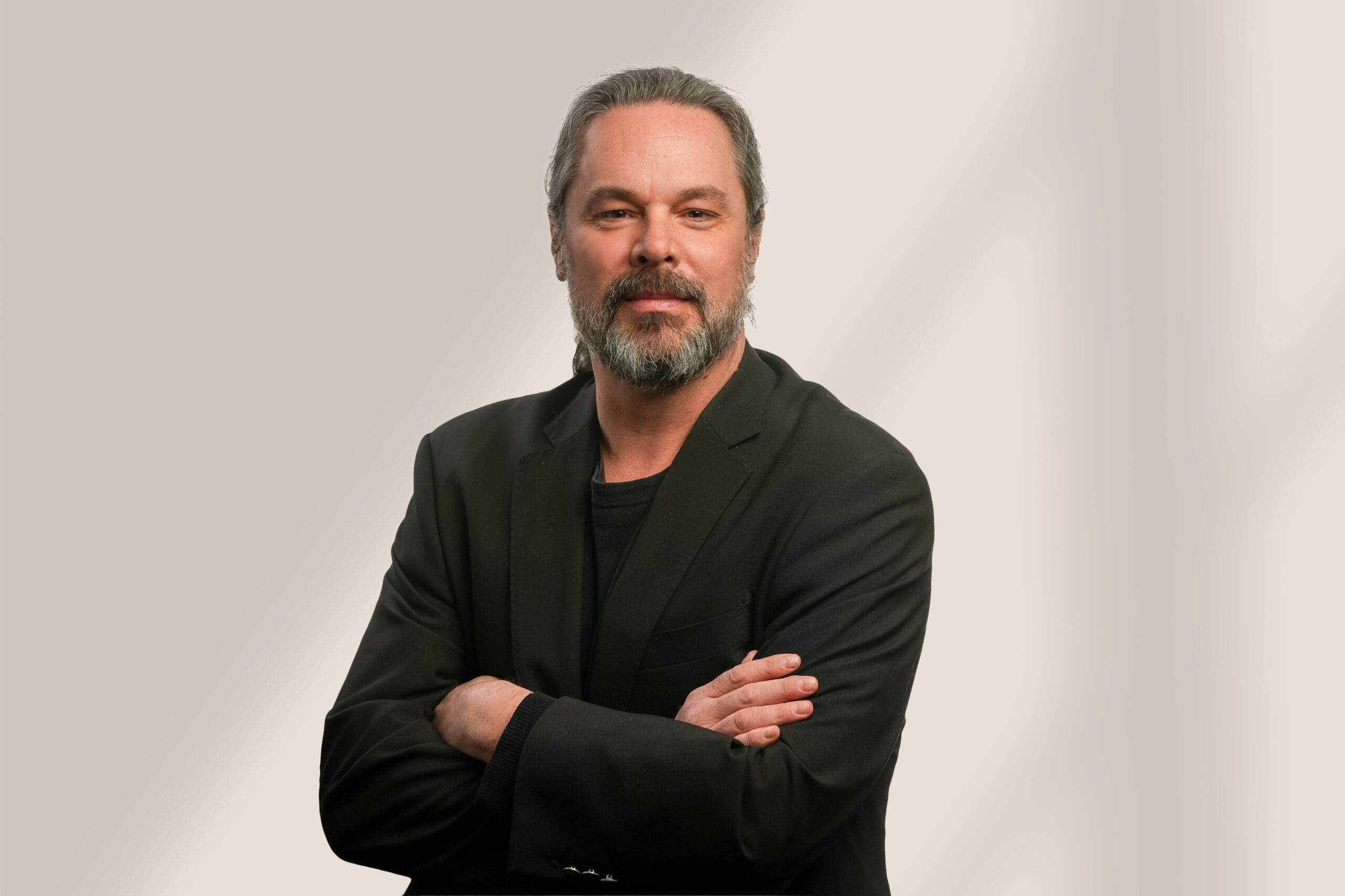 Un homme d'âge moyen à la barbe grise et aux cheveux longs, vêtu d'un blazer et d'une chemise noirs, se tient debout, les bras croisés, sur un fond clair et neutre.
