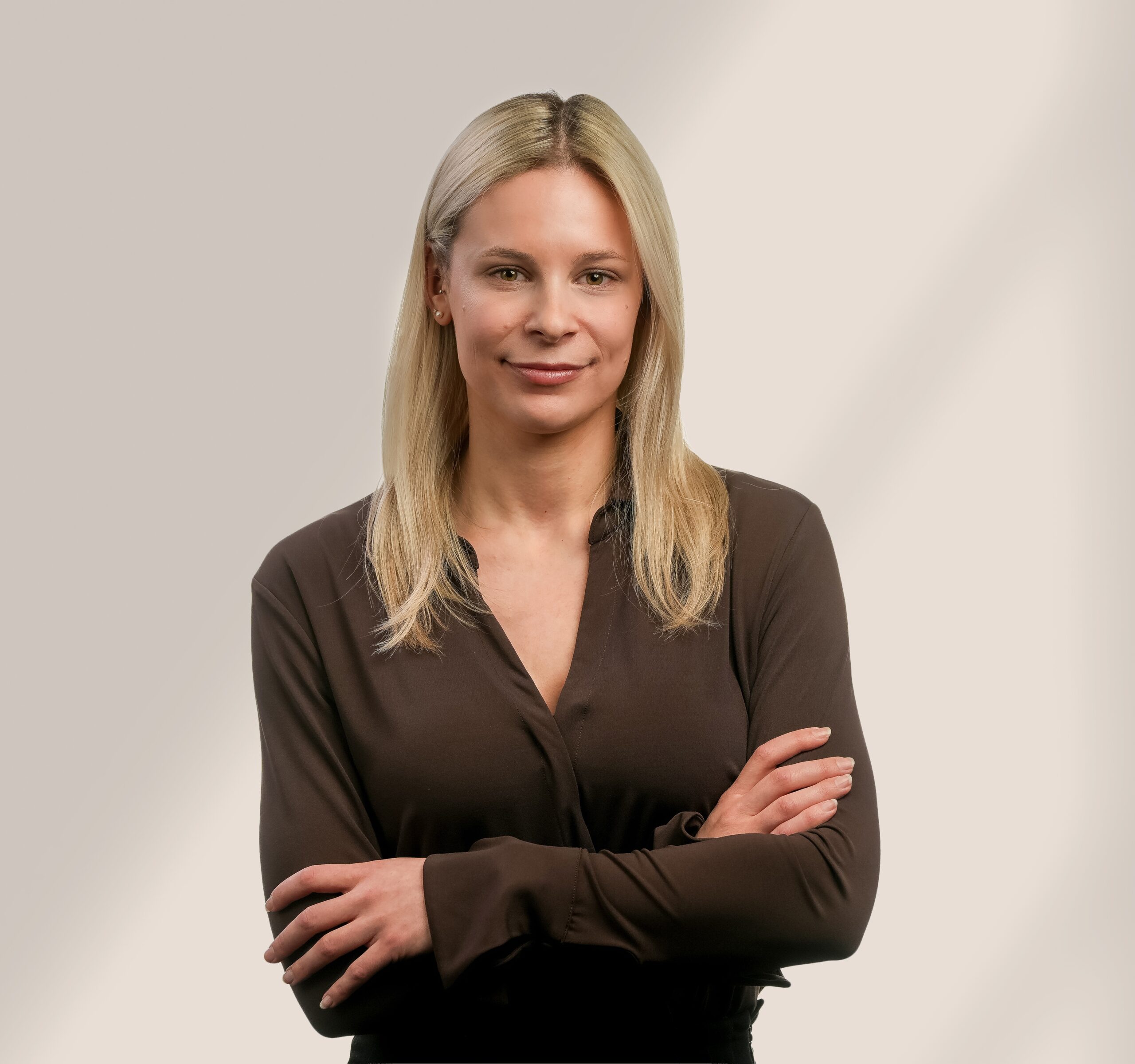 Une femme aux cheveux blonds et raides, vêtue d'un chemisier sombre à manches longues, se tient sur un fond clair et uni, les bras croisés, souriant doucement à l'appareil photo.