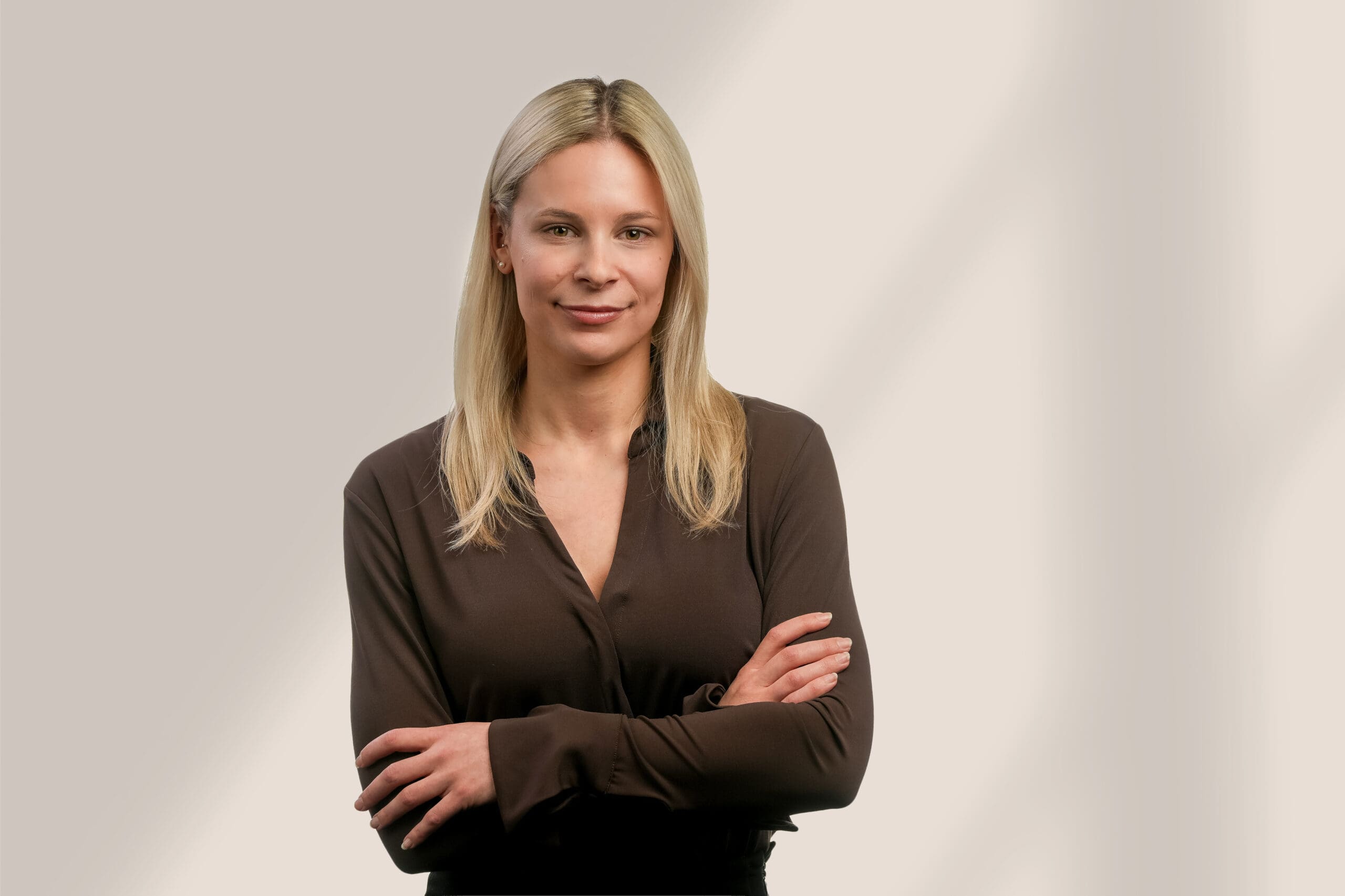 Une femme aux cheveux blonds et raides, vêtue d'un chemisier sombre à manches longues, se tient debout, les bras croisés et sourit légèrement sur un fond uni de couleur claire.