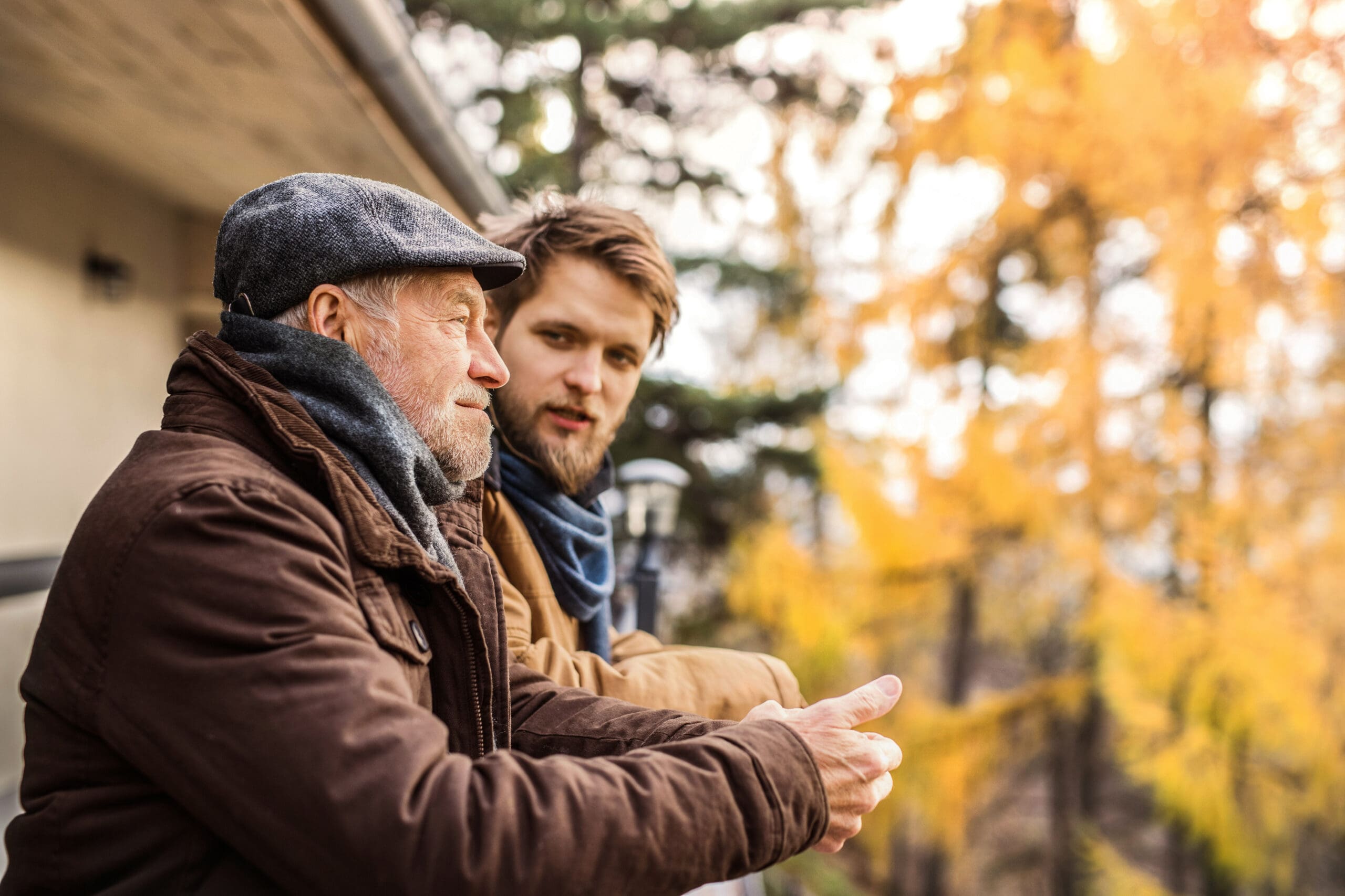 Deux hommes, l'un plus âgé et l'autre plus jeune, portent des manteaux et des écharpes et se tiennent sur un balcon, en pleine conversation. Des arbres d'automne aux feuilles jaunes se trouvent à l'arrière-plan.