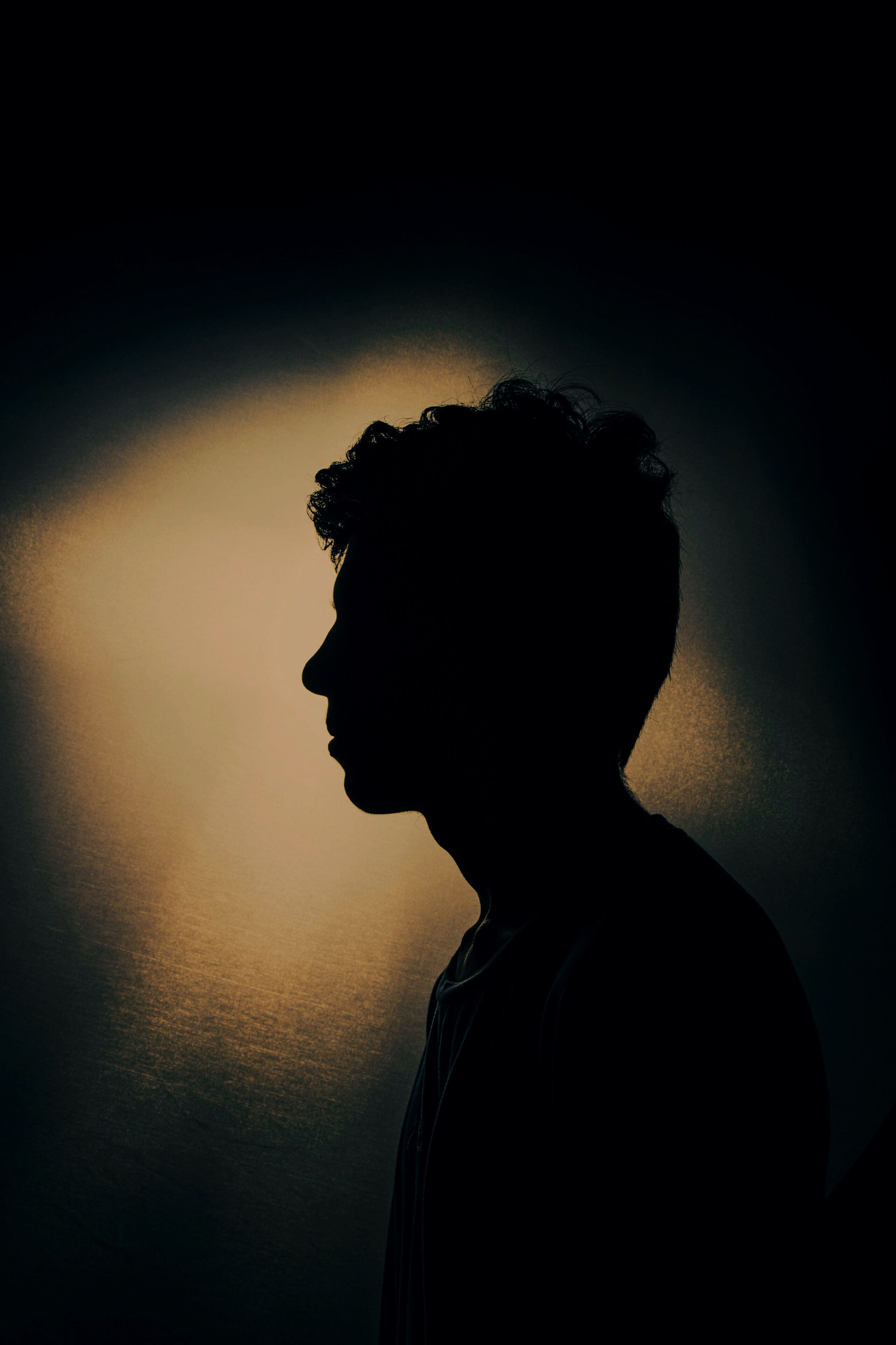 Silhouette d'une personne de profil tournée vers la gauche, aux cheveux bouclés, sur un fond sombre et une lumière douce créant un effet de halo derrière sa tête.