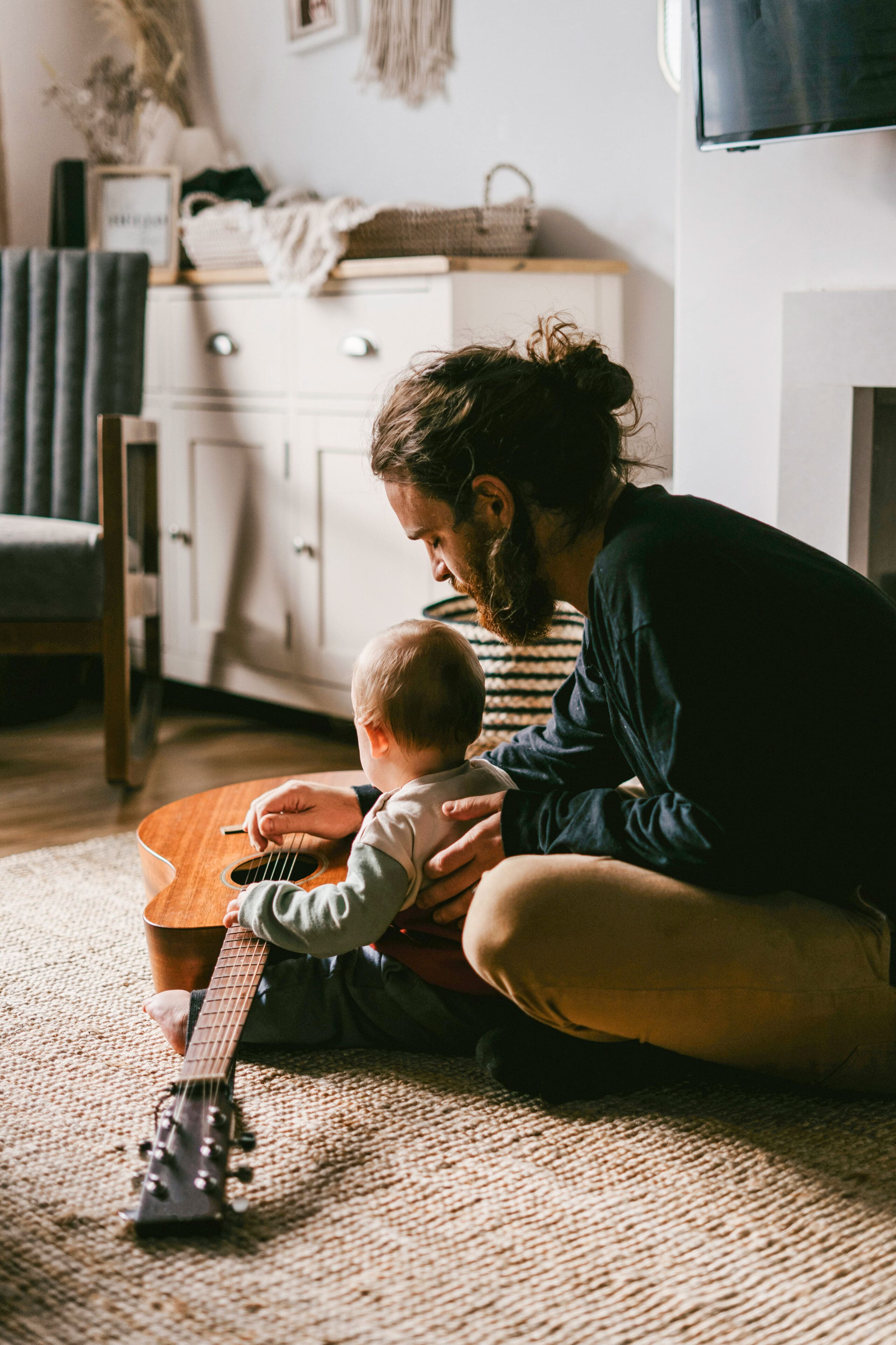 Un homme barbu est assis par terre avec un bébé, guidant la main de l'enfant sur une guitare acoustique. Ils se trouvent dans un salon confortable, légèrement éclairé, avec un tapis et des meubles en arrière-plan.